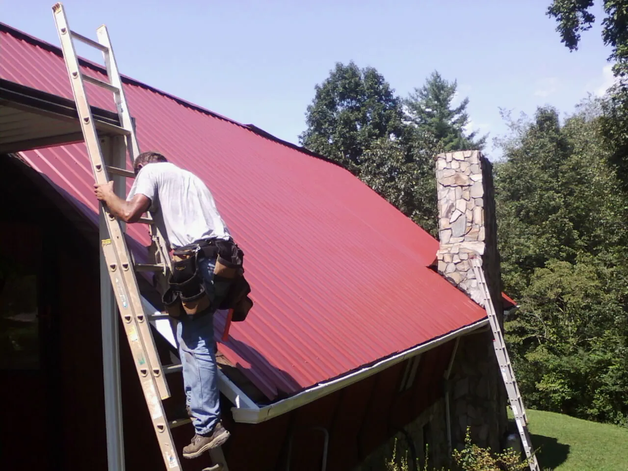 Roofer on a ladder beside a red metal roof and stone chimney — Soffit & Fascia Repair by Roofing Brothers in Glenmoore